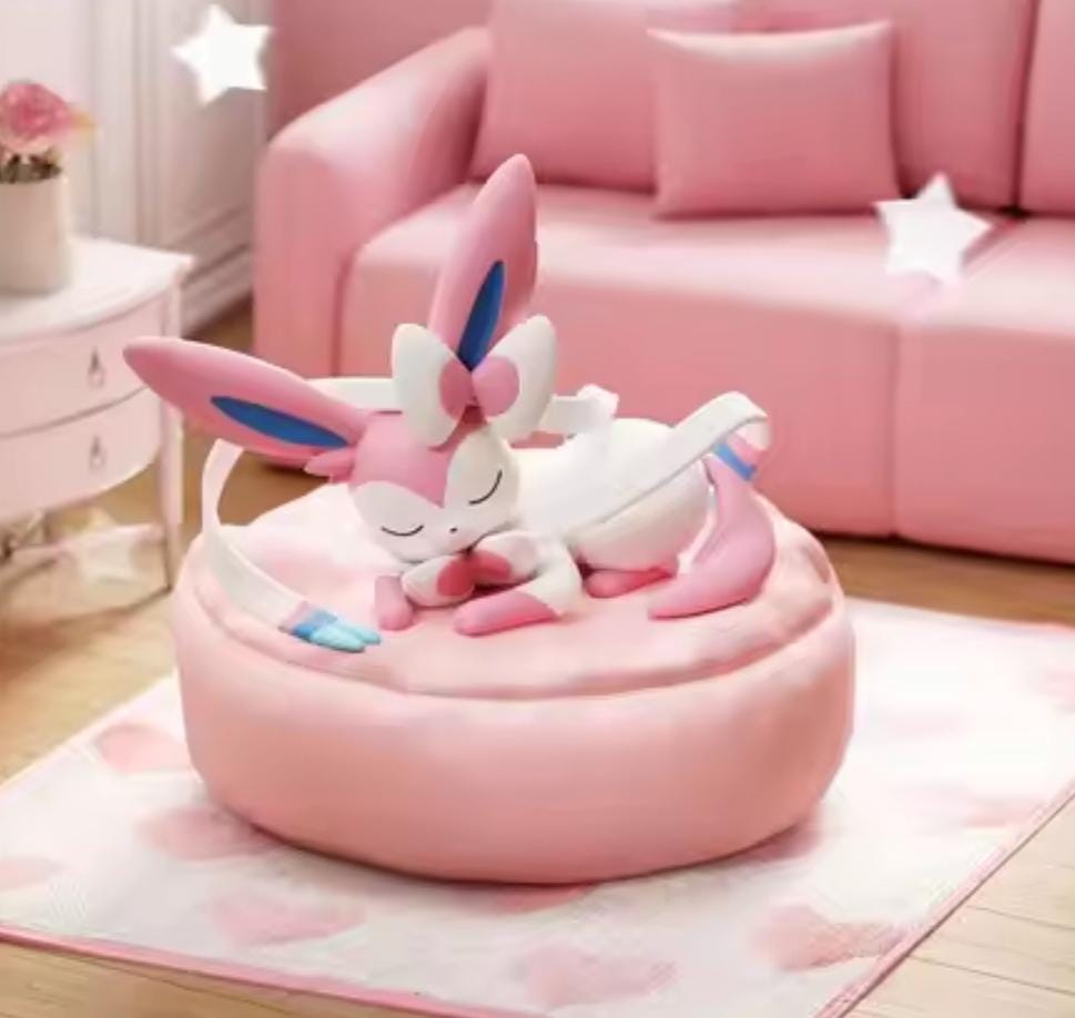 Close-up of sleeping Sylveon figure with blue ribbon details resting on a large pink soft-touch cushion, part of the Dreamy Evolution Collection.